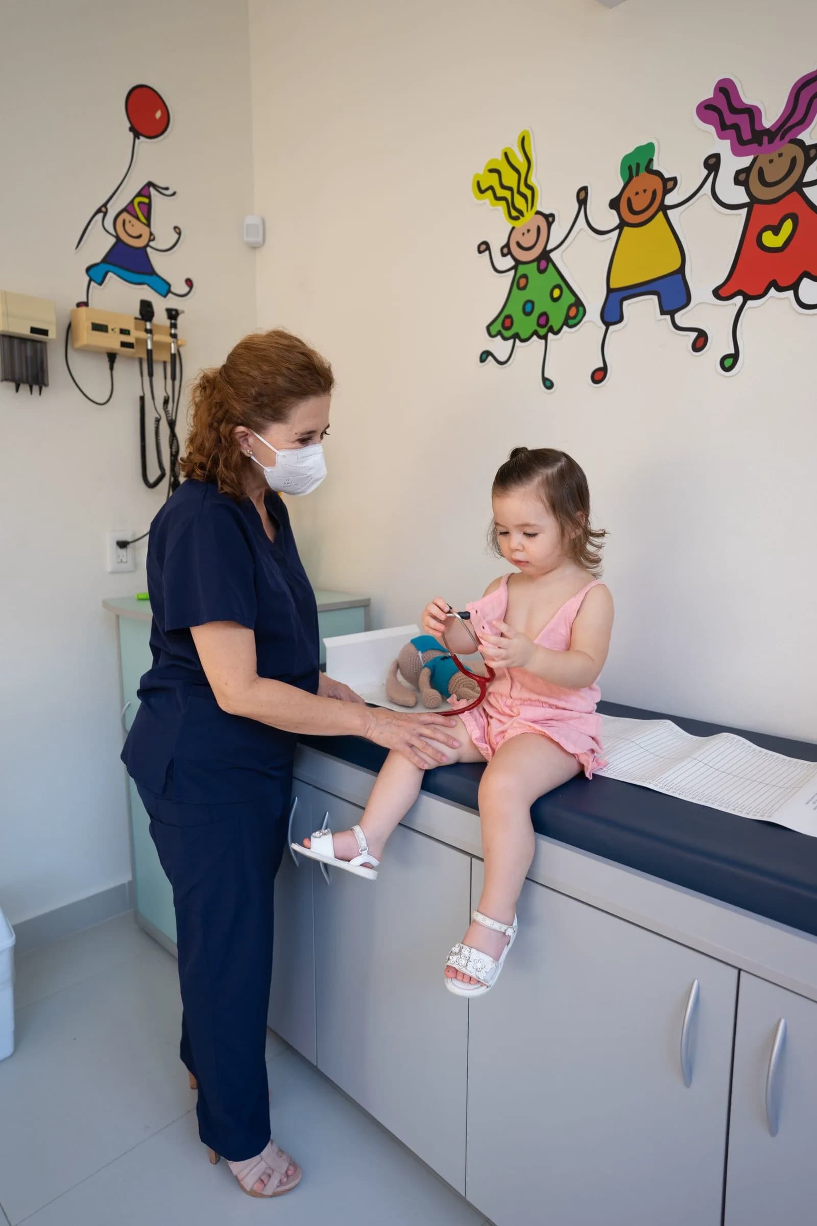 Pediatrician standing with a young child in a clinic environment, parents present in the background
