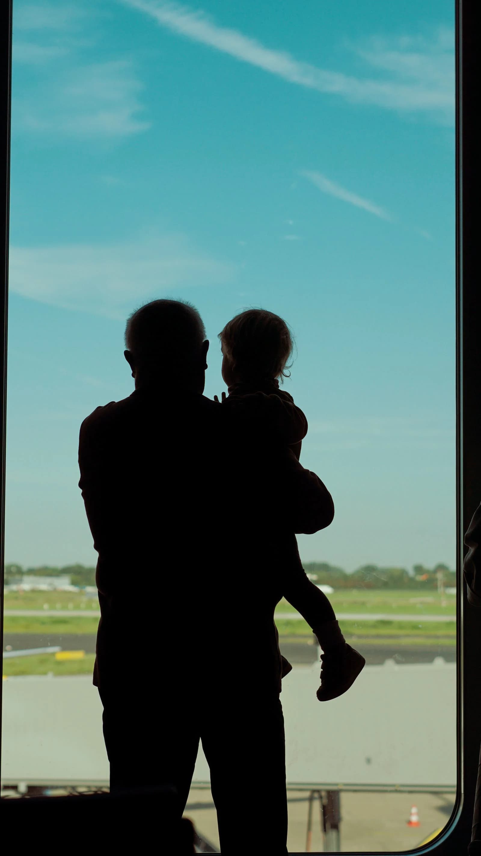 Silhouette of a parent holding a child at a large airport window overlooking the runway