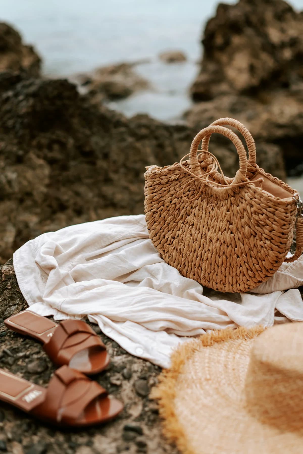 Beach accessories including straw hat, sandals, and tote bag arranged on a sunny rock