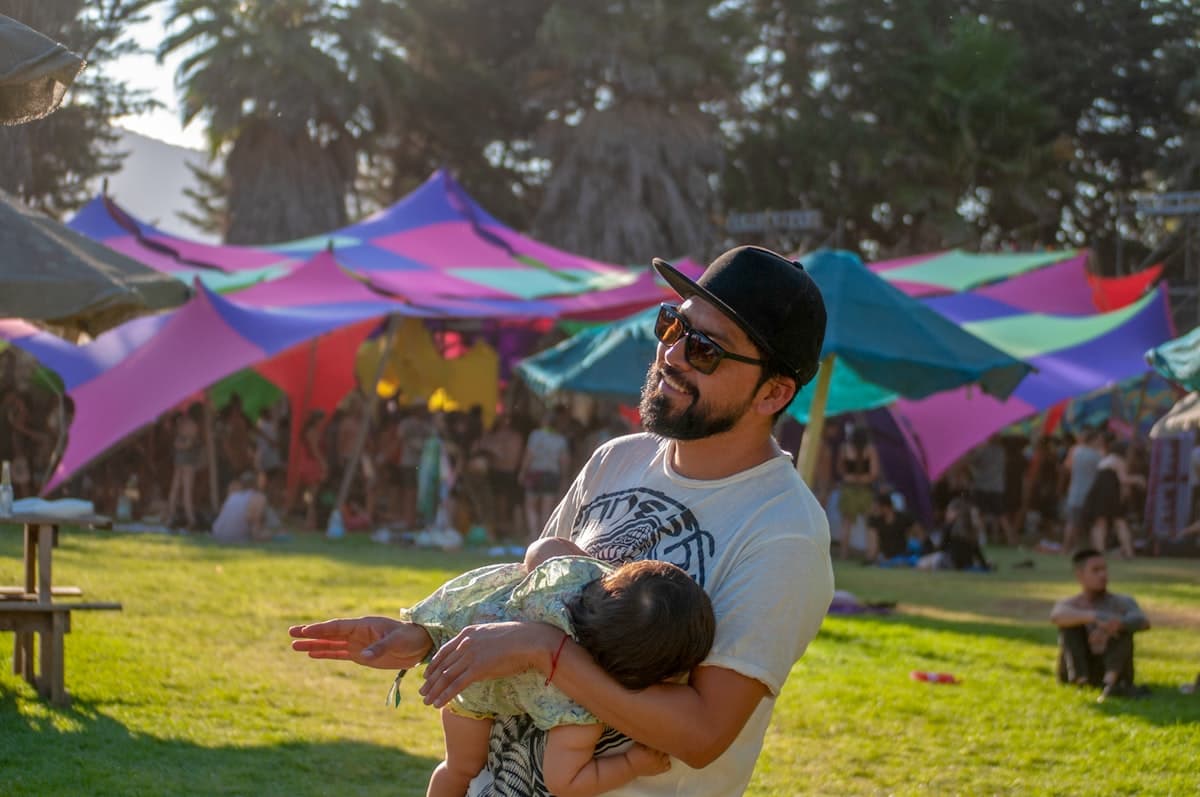 Parent carrying baby near a shaded canopy at a summer outdoor event